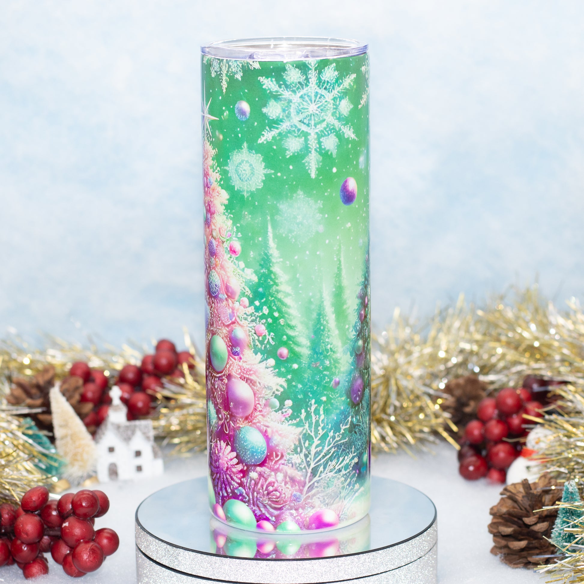 A tall cylindrical tumbler with a clear lid, featuring a festive Christmas tree design in shades of green, pink, purple, and blue. The tumbler is displayed on a silver pedestal with holiday decorations like red berries, pinecones, and gold tinsel in the background.
