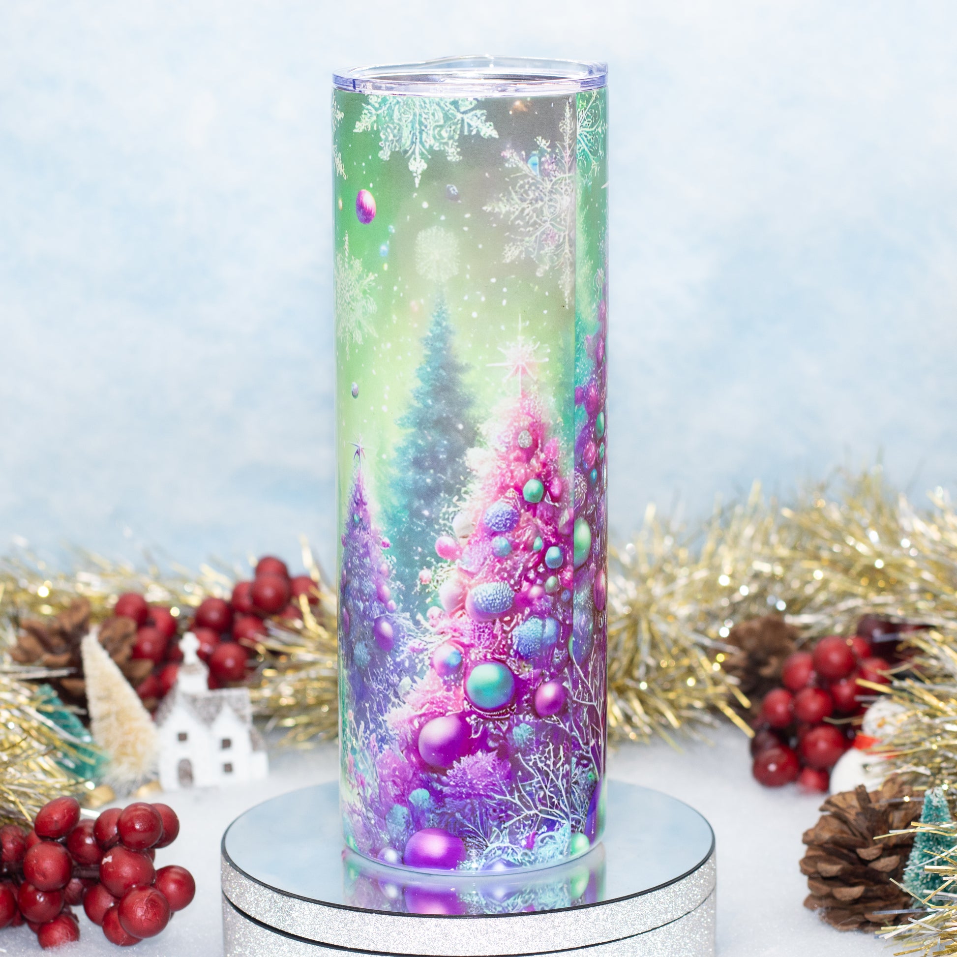 A cylindrical tumbler with a festive Christmas scene printed on it, featuring purple and green Christmas trees with blue ornaments, displayed on a silver glitter stand with holiday decorations in the background.