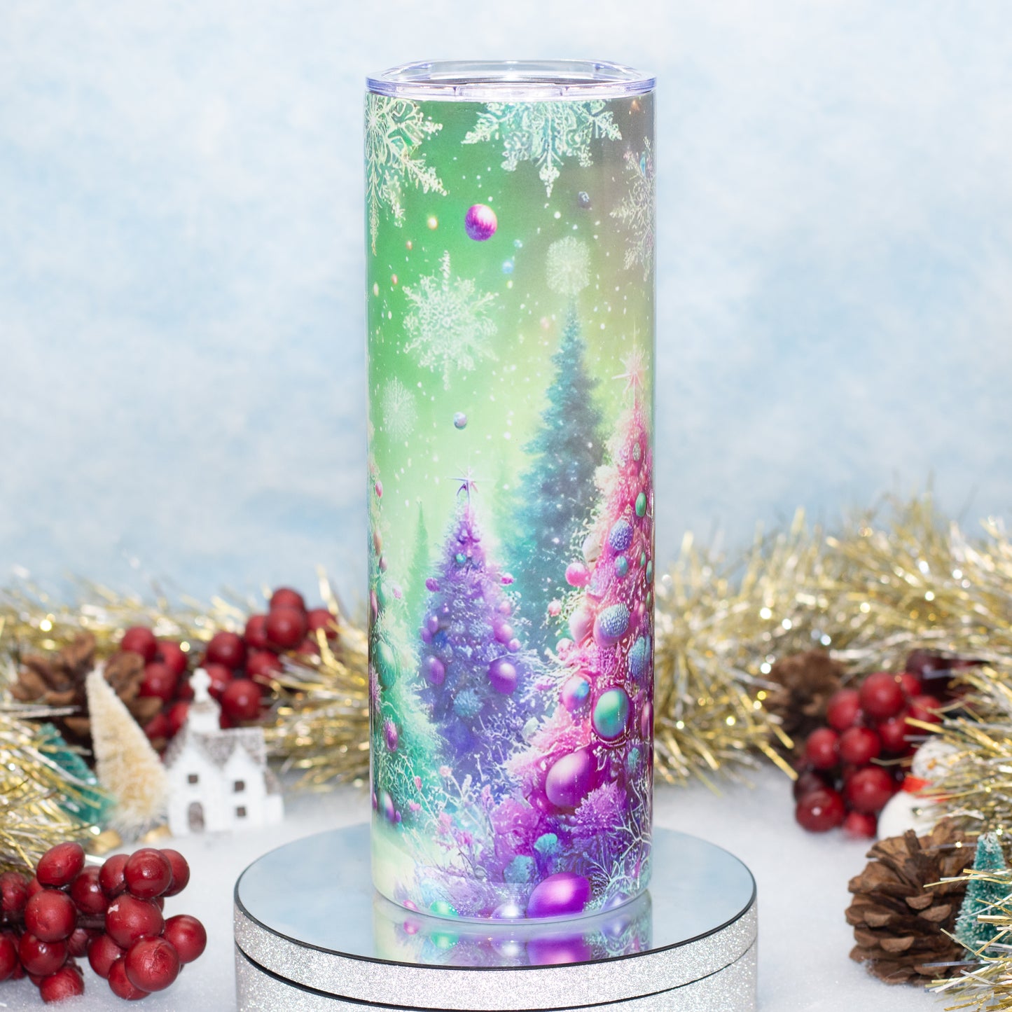 A tall cylindrical tumbler with a clear lid, featuring a colorful winter forest design with purple and green Christmas trees, snowflakes, and ornaments. The tumbler is displayed on a silver glitter pedestal with holiday decorations like red berries, pinecones, and gold tinsel in the background.