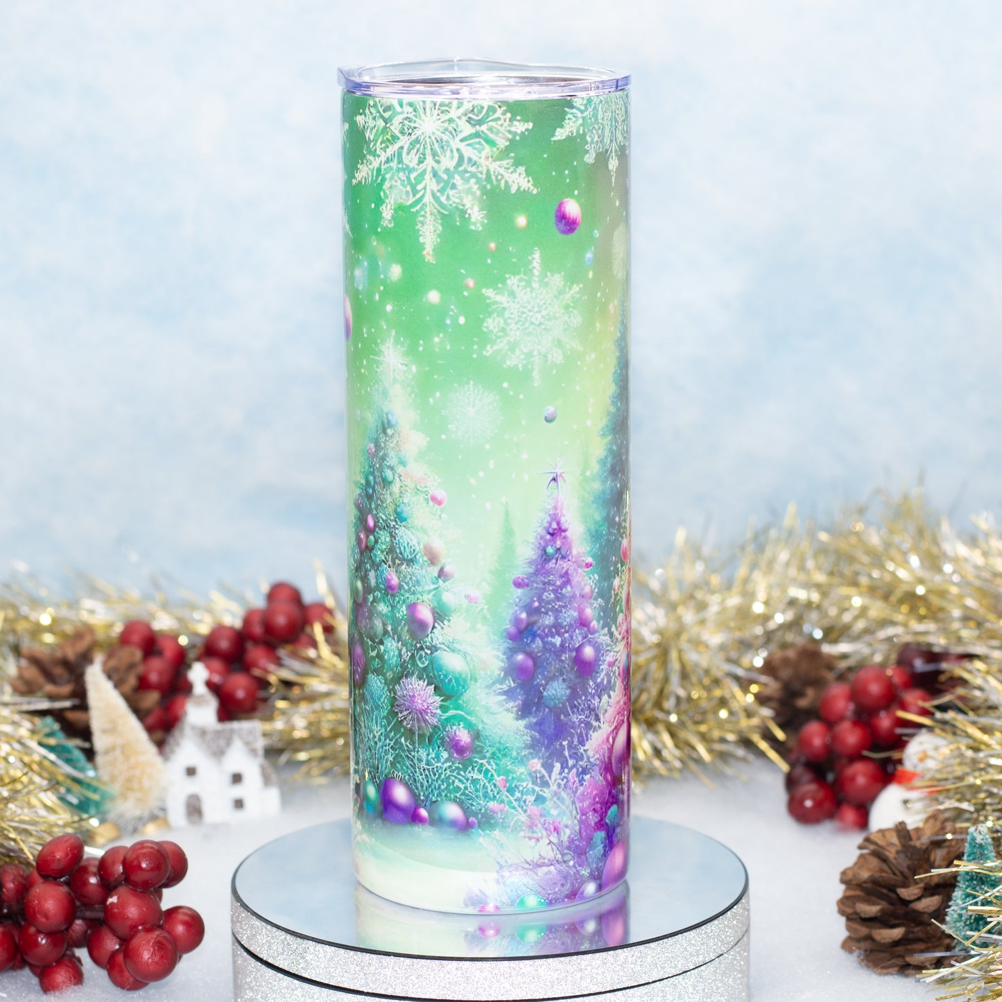 A tall cylindrical tumbler with a festive Christmas winter scene print, featuring snowflakes, evergreen trees, and purple ornaments, displayed on a silver round pedestal with holiday decorations in the background.