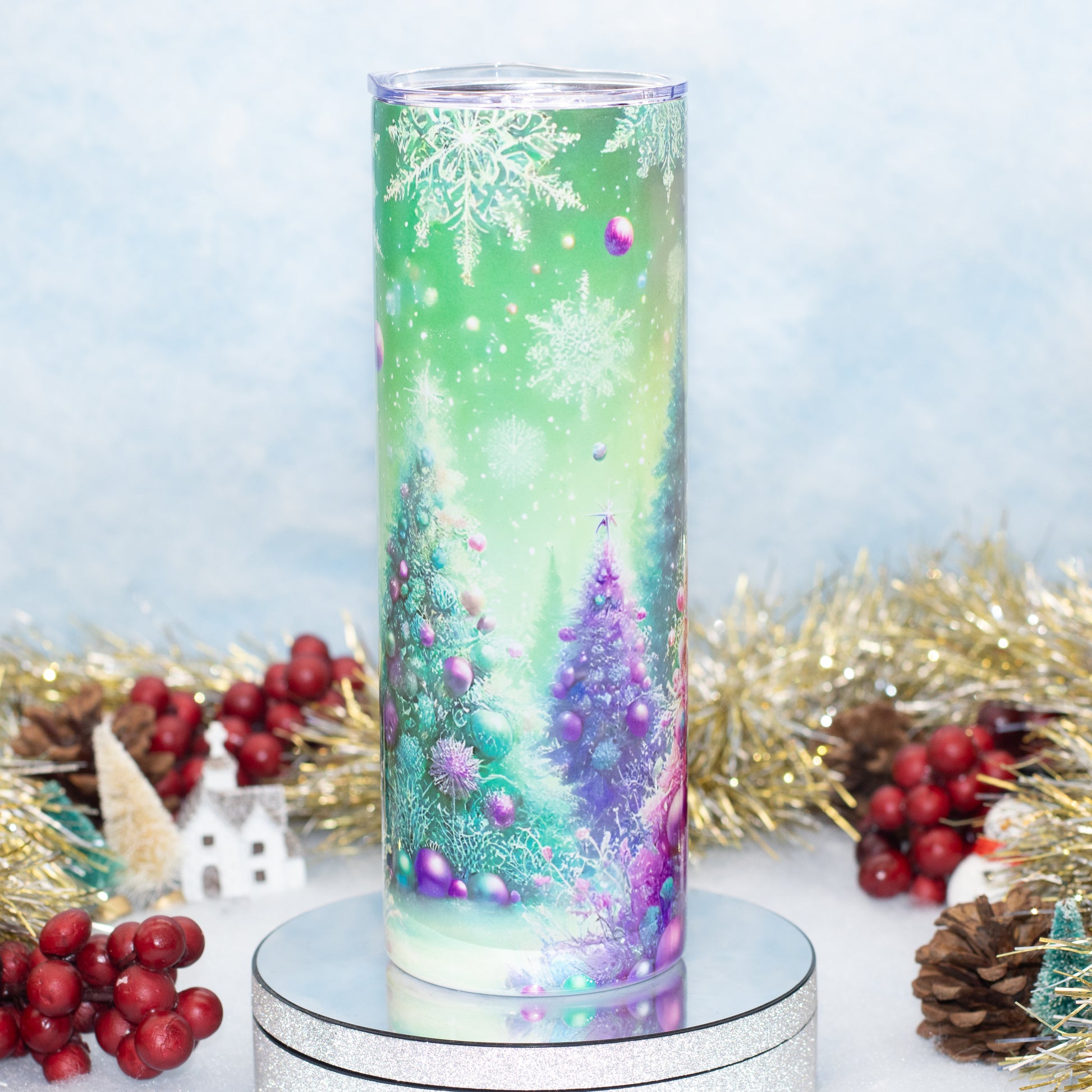 A tall cylindrical tumbler with a festive Christmas winter scene print, featuring snowflakes, evergreen trees, and purple ornaments, displayed on a silver round pedestal with holiday decorations in the background.