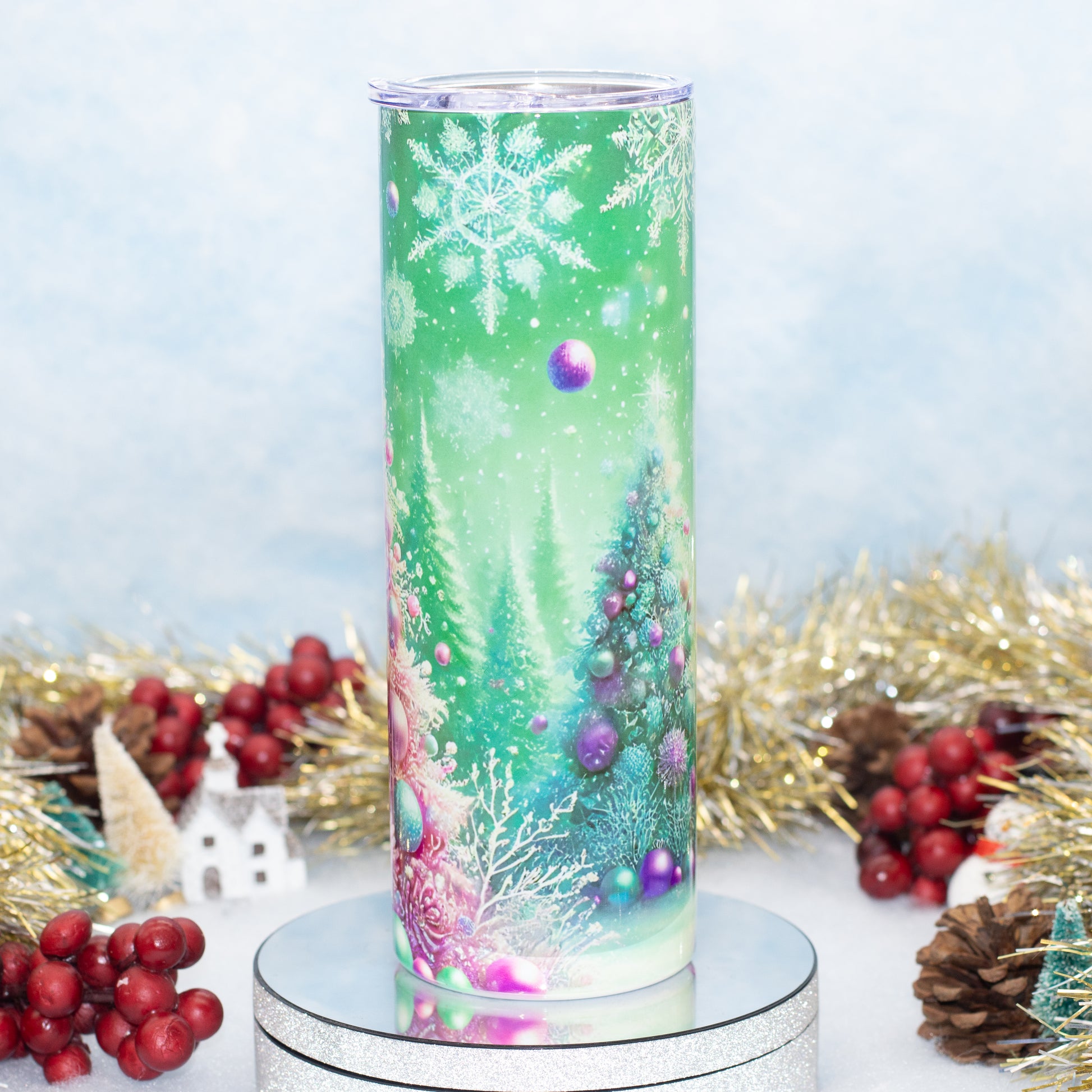 A tall, skinny tumbler with a festive winter holiday design, featuring snowflakes, Christmas trees, and purple ornaments on a green gradient background, displayed on a silver pedestal with holiday decorations.
