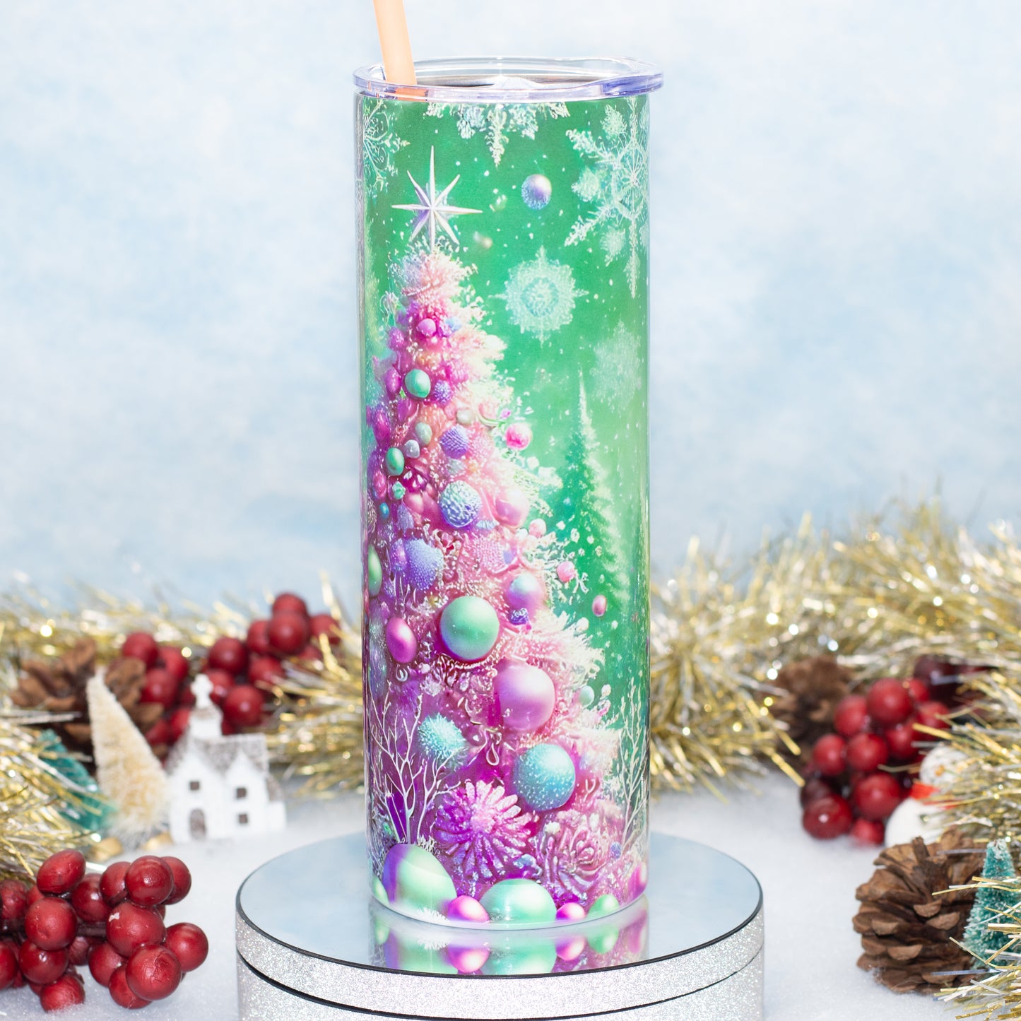 A tall clear tumbler with a printed Christmas tree design in green, pink, purple, and blue, shown on a silver pedestal with holiday decorations around it.
