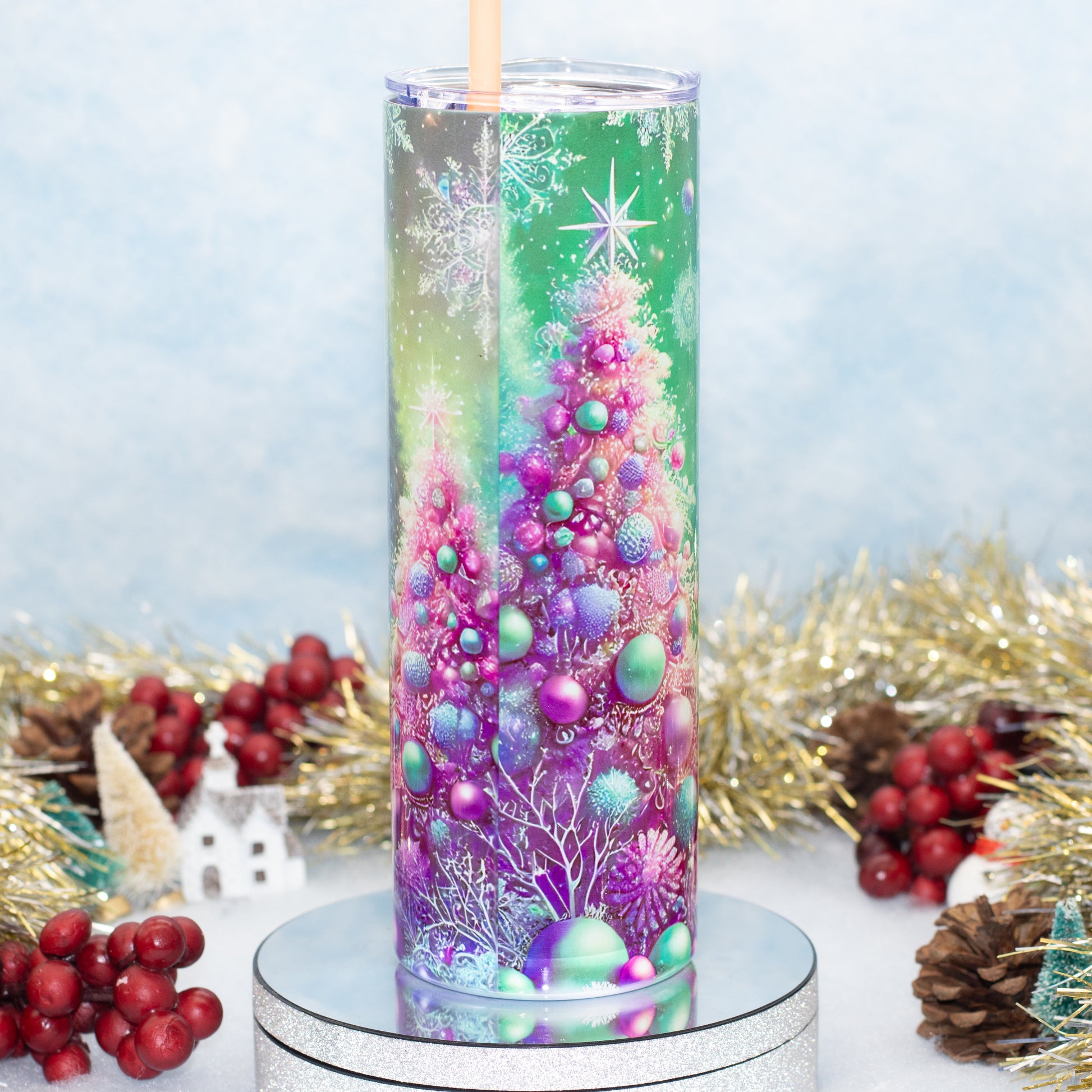 A tall clear tumbler with a straw, decorated with a colorful Christmas tree design in purple, pink, blue, and green, displayed on a silver pedestal with holiday decorations in the background.