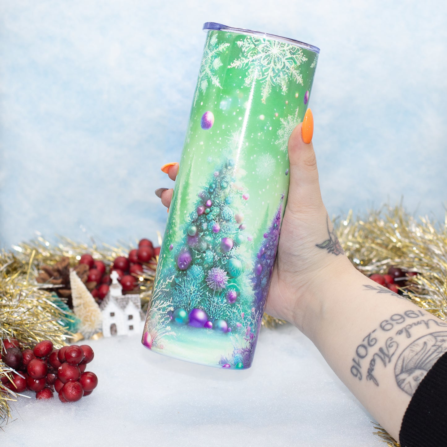 A hand holds a tall insulated tumbler with a festive Christmas tree design. The tumbler features a green and blue winter tree illustration with purple ornaments and snowflakes.