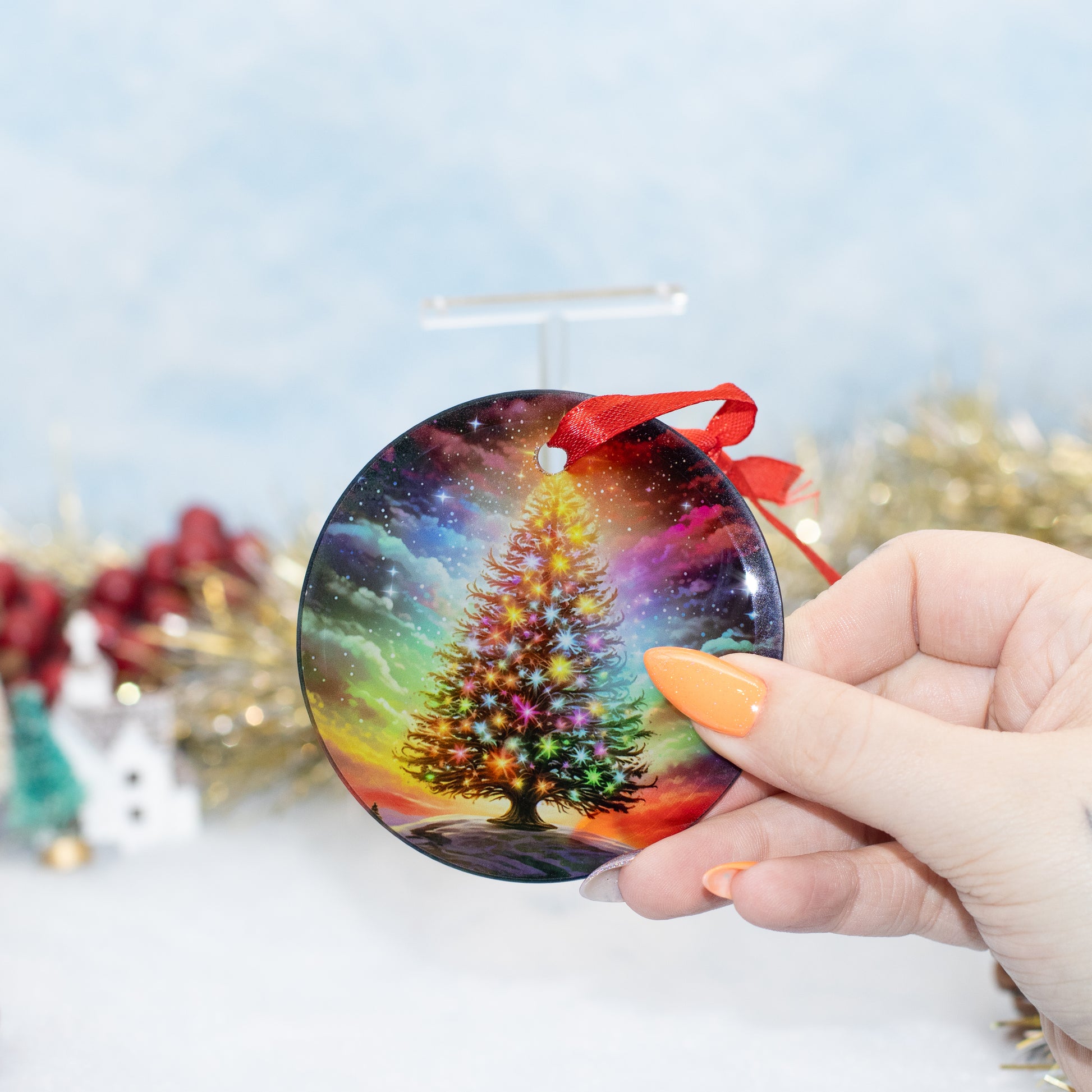 A hand holds a round Christmas ornament featuring a colourful, glowing Christmas tree against a starry galaxy sky. The ornament has a red ribbon loop and is shown against a festive, blurred holiday background.