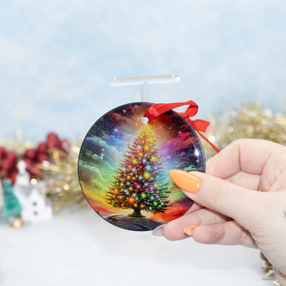 A hand holds a round Christmas ornament featuring a colourful, glowing Christmas tree against a starry galaxy sky. The ornament has a red ribbon loop and is shown against a festive, blurred holiday background.