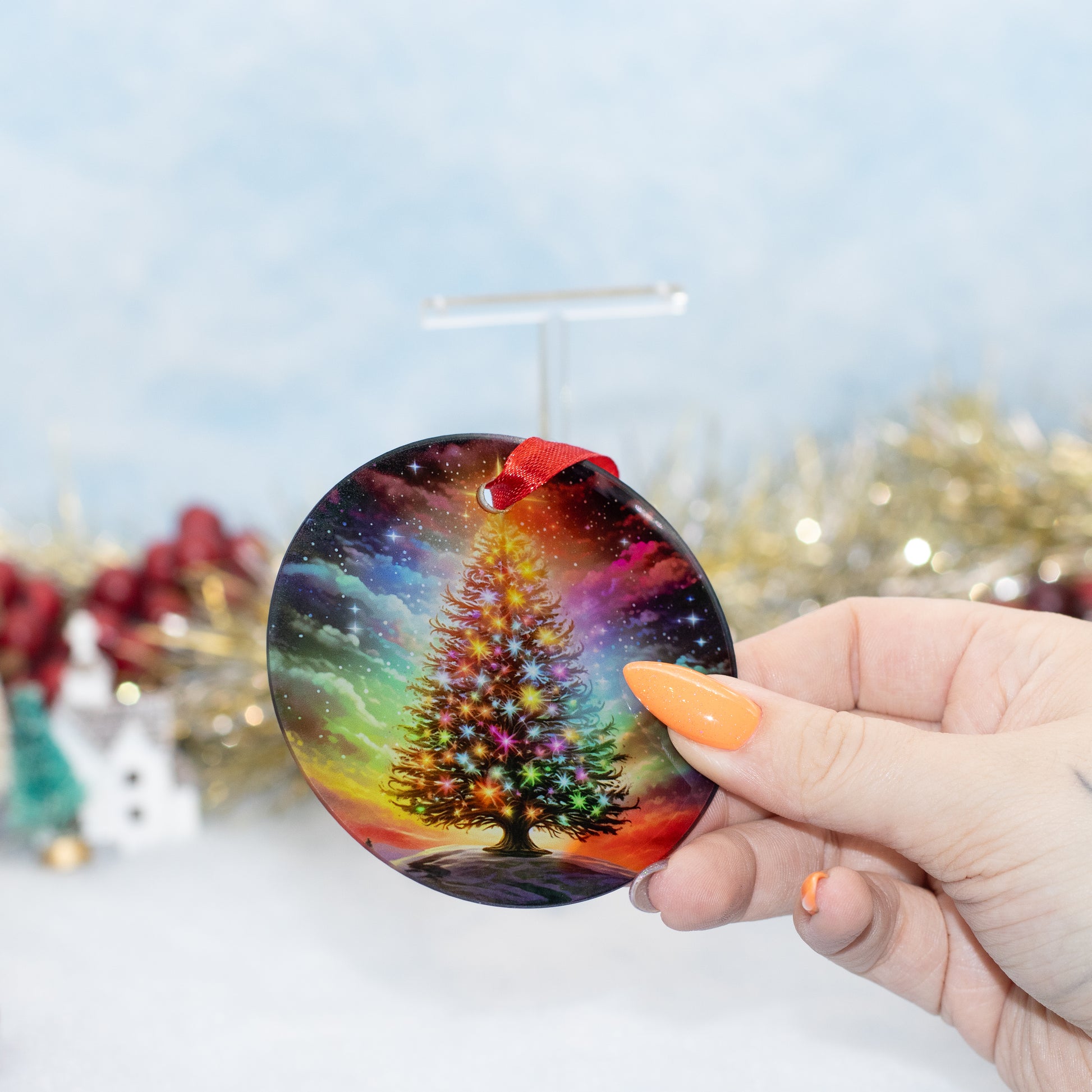 A hand holds a round Christmas ornament featuring a colourful, glowing Christmas tree under a starry night sky. The background is a soft-focus winter scene with tinsel and snow.