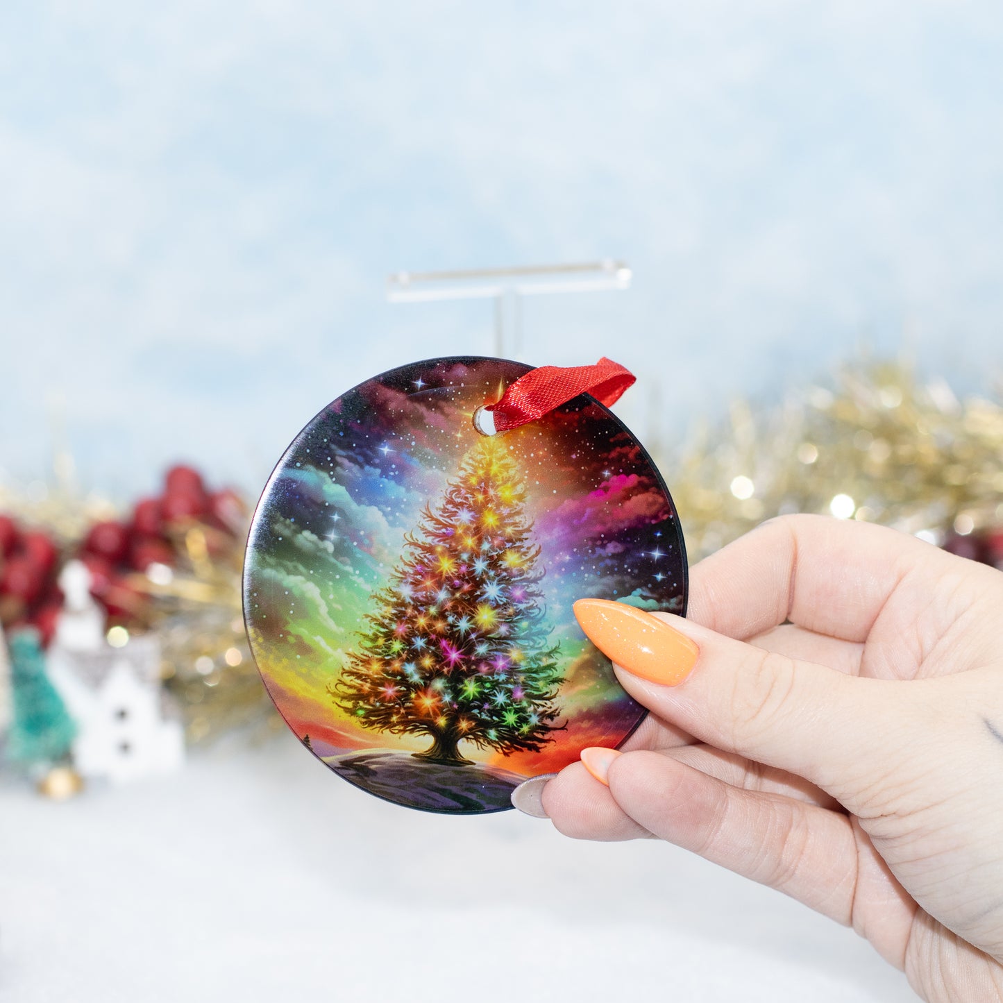 A hand holds a round ceramic Christmas ornament featuring a colourful, glowing Christmas tree against a starry space background. The ornament has a red hanging ribbon and is photographed against a soft, out-of-focus holiday backdrop with tinsel and miniature decorations.