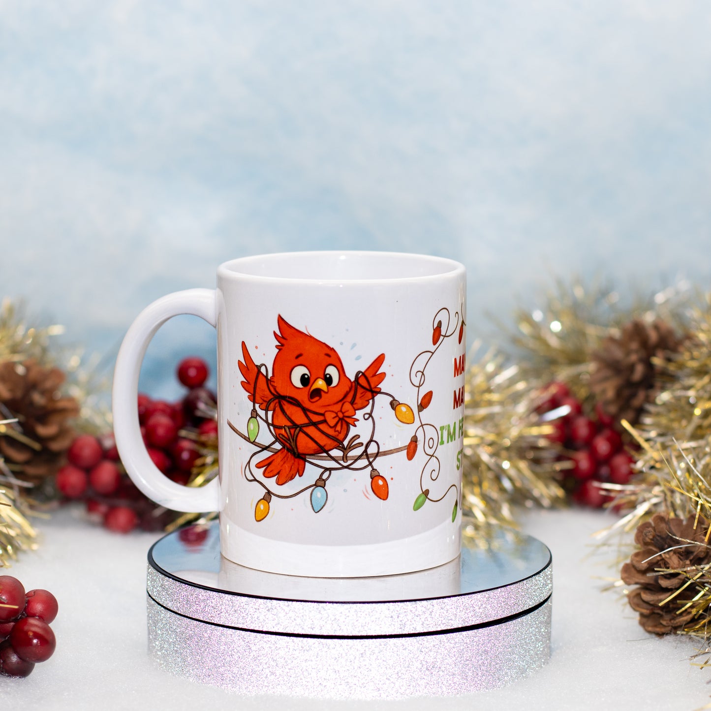 A white ceramic mug featuring a festive Christmas design of a red cardinal surrounded by colorful holiday lights, displayed on a round silver glitter stand with holiday decorations in the background.