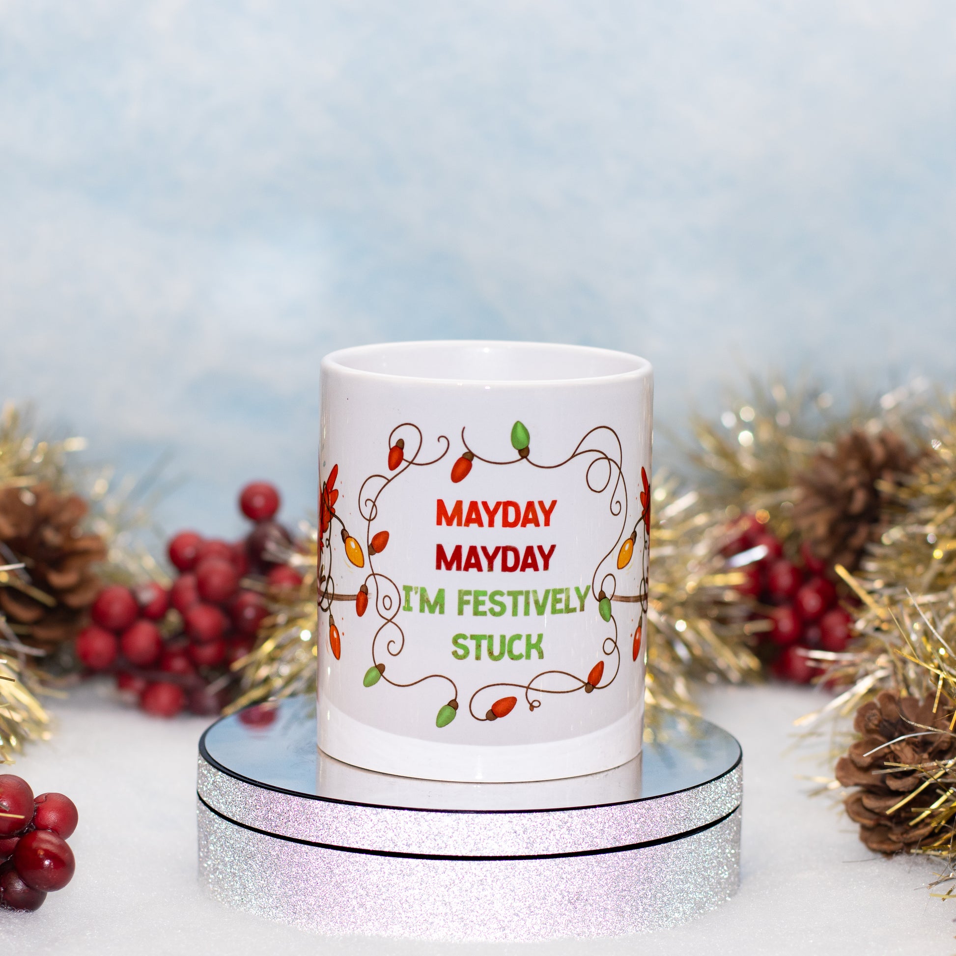 A white ceramic mug with a festive holiday light design and the text 'MAYDAY MAYDAY I'M FESTIVELY STUCK' in red and green lettering. The mug is displayed on a round silver mirror stand, surrounded by holiday decorations 