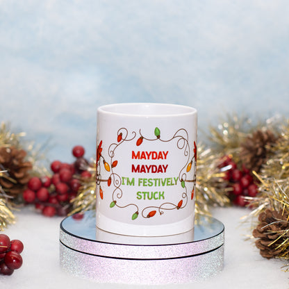 A white ceramic mug with a festive holiday light design and the text 'MAYDAY MAYDAY I'M FESTIVELY STUCK' in red and green lettering. The mug is displayed on a round silver mirror stand, surrounded by holiday decorations 