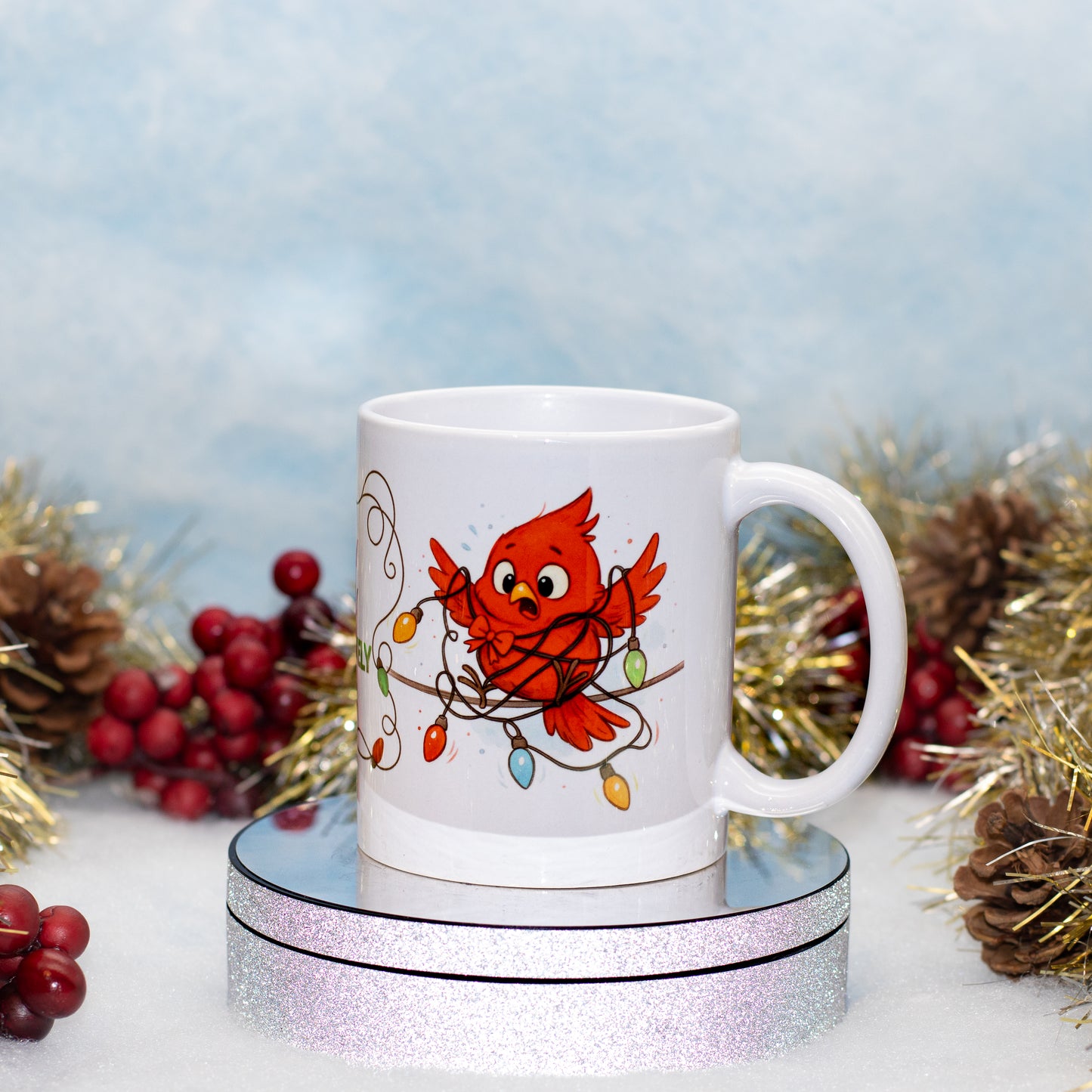 A white ceramic mug featuring a red cardinal wrapped in holiday string lights, displayed on a round silver glitter pedestal with festive tinsel and berries in the background.