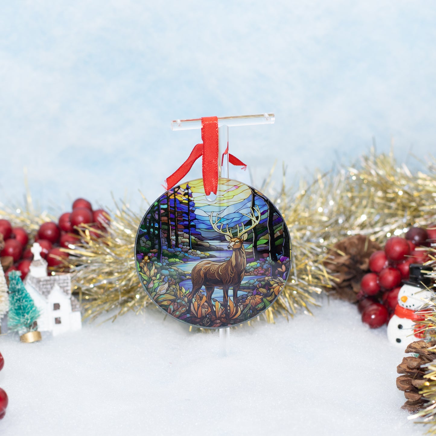A round Christmas ornament featuring a painted deer in a forest under a full moon, hanging from a red ribbon. The ornament is styled on a white surface with gold tinsel, red berries, a small tree, and a snowman decoration against a soft light blue background.