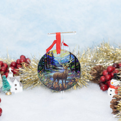 A round Christmas ornament featuring a painted deer in a forest under a full moon, hanging from a red ribbon. The ornament is styled on a white surface with gold tinsel, red berries, a small tree, and a snowman decoration against a soft light blue background.