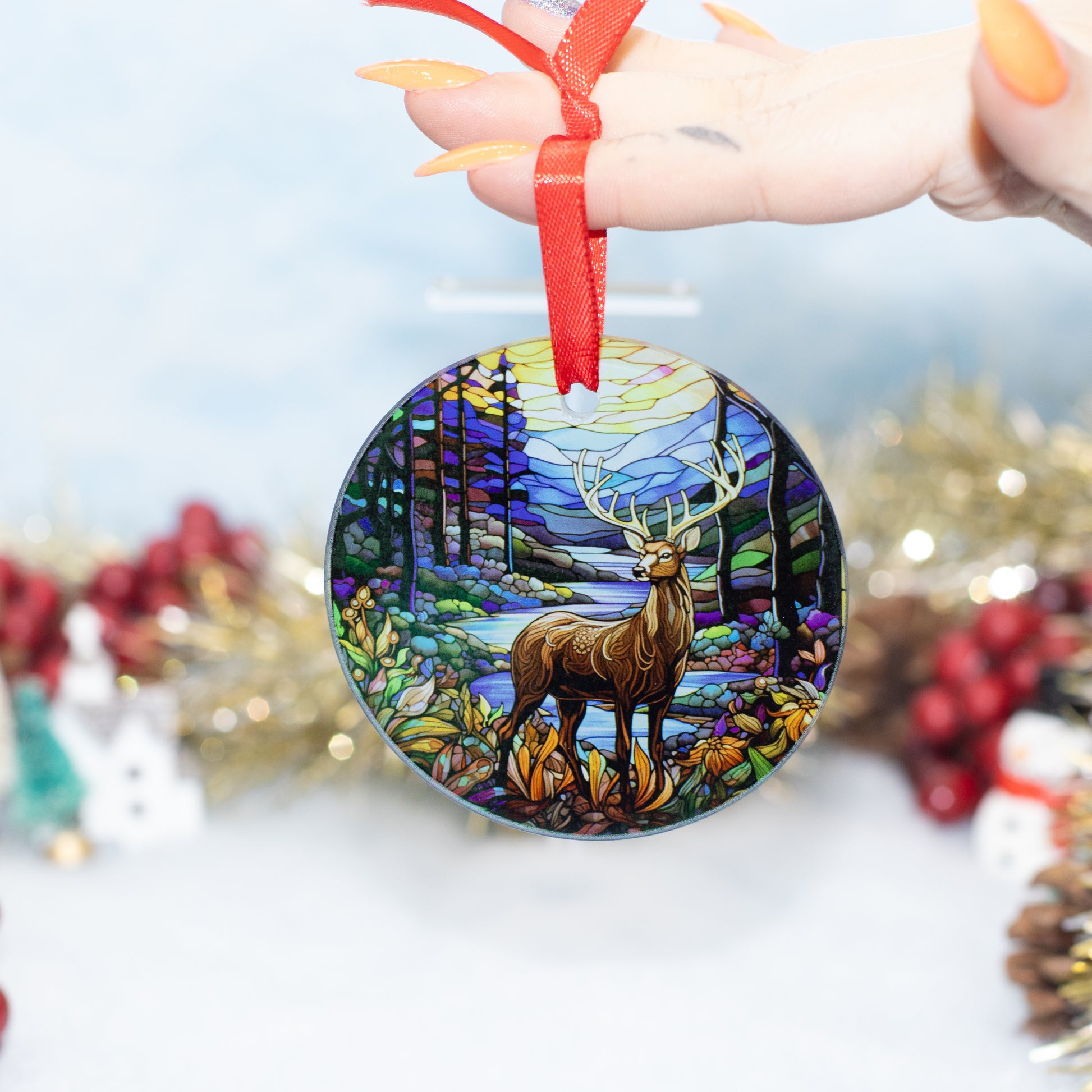 A hand holds a round Christmas ornament featuring a stained-glass-style forest scene with a deer, suspended by a red ribbon against a softly blurred festive background.