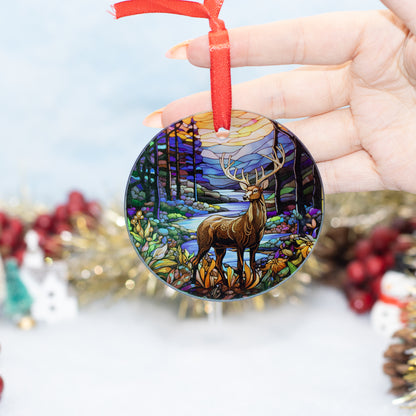 A hand holds a round Christmas ornament featuring a stained-glass style winter forest scene with a deer and blue stream. The ornament has a red ribbon hanger and is photographed against a softly blurred festive background.