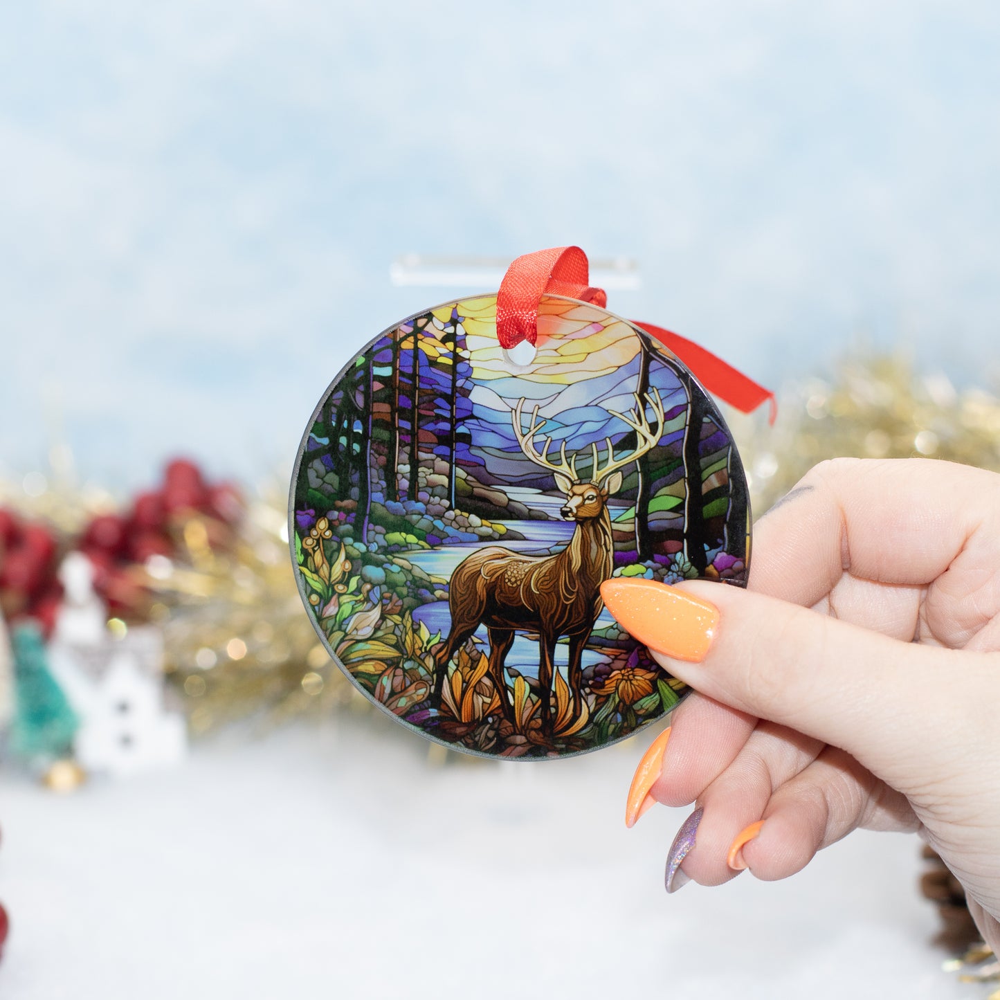A hand holds a round stained-glass style Christmas ornament featuring a deer in a colourful forest. The ornament has a red ribbon loop and is photographed against a softly blurred festive background.