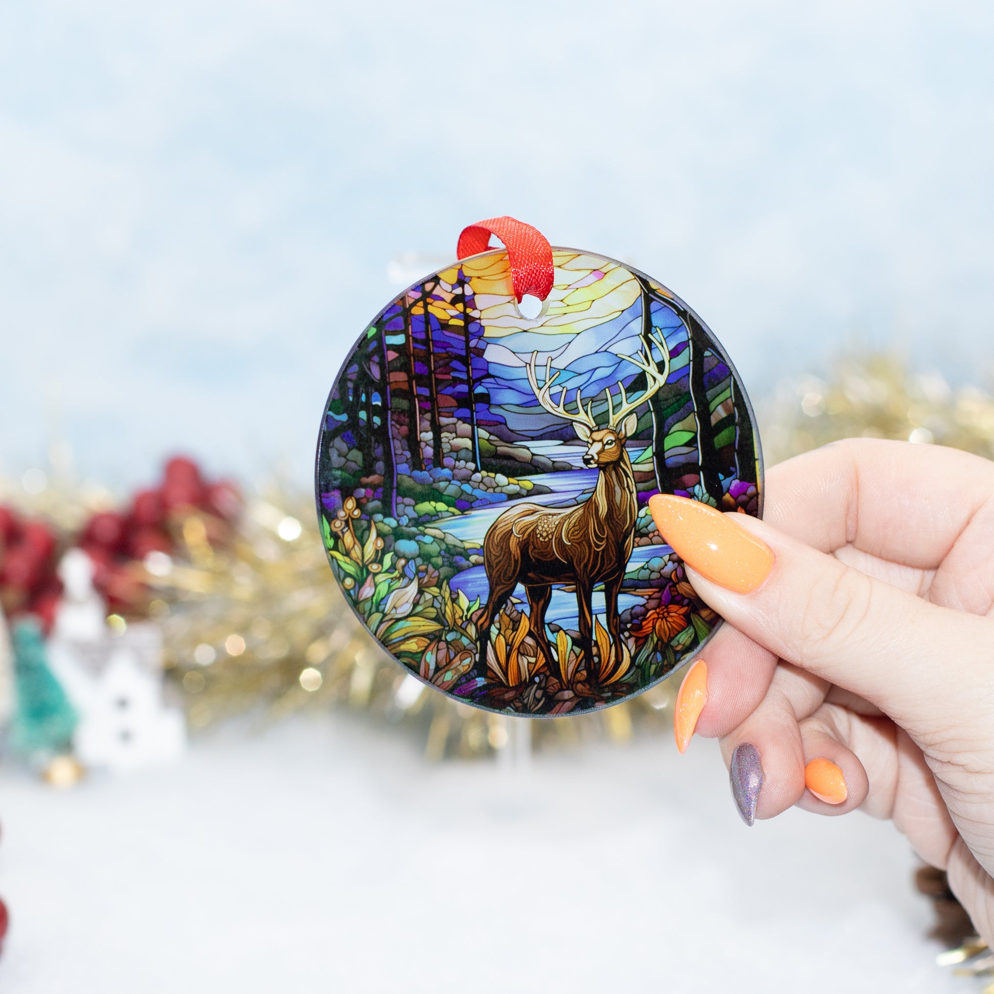 A hand holds a round stained-glass-style Christmas ornament featuring a deer in a colourful forest. The ornament has a red hanging loop and is shown against a softly blurred holiday background with tinsel and ornaments.