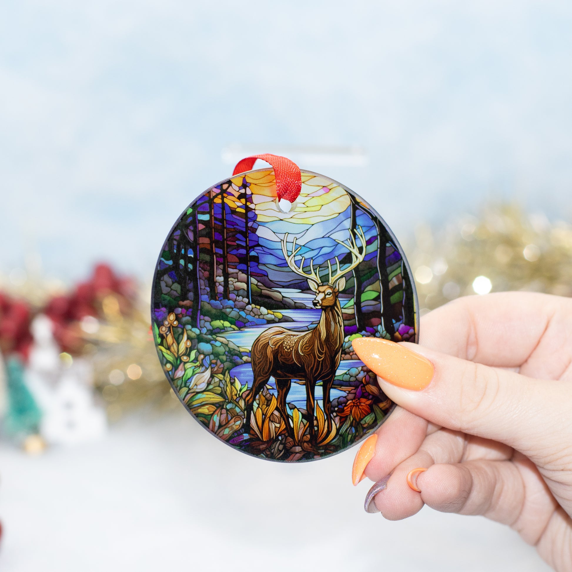 A hand holds a round stained-glass-style holiday ornament featuring a deer in a colourful forest landscape. The ornament has a red hanging ribbon and is photographed against a softly blurred festive background.
