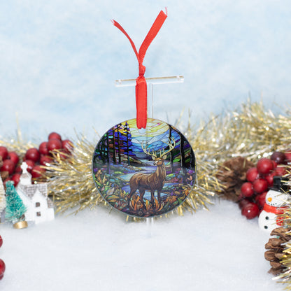 A round Christmas ornament featuring a painted forest scene with a deer, hanging from a red ribbon on a clear stand. The ornament is styled on a snowy surface with gold tinsel, red berries, pinecones, and small holiday decorations in the background.