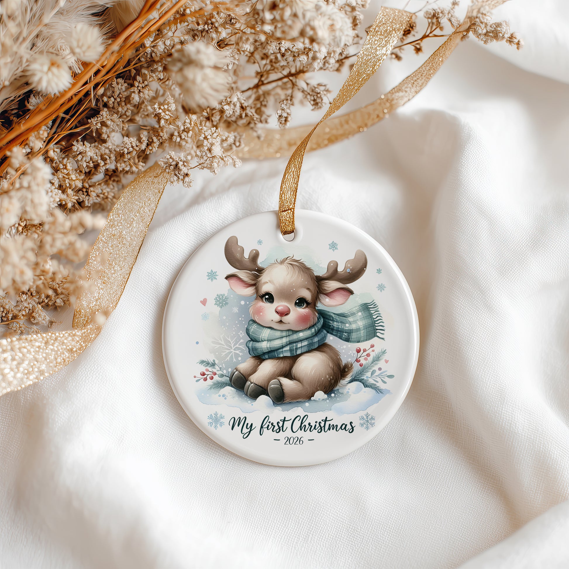 A round white ceramic Christmas ornament featuring a watercolor illustration of a baby deer wearing a blue scarf, with the text 'My first Christmas 2026' printed below. The ornament hangs from a gold ribbon and is styled on white fabric with dried flowers and a gold ribbon.