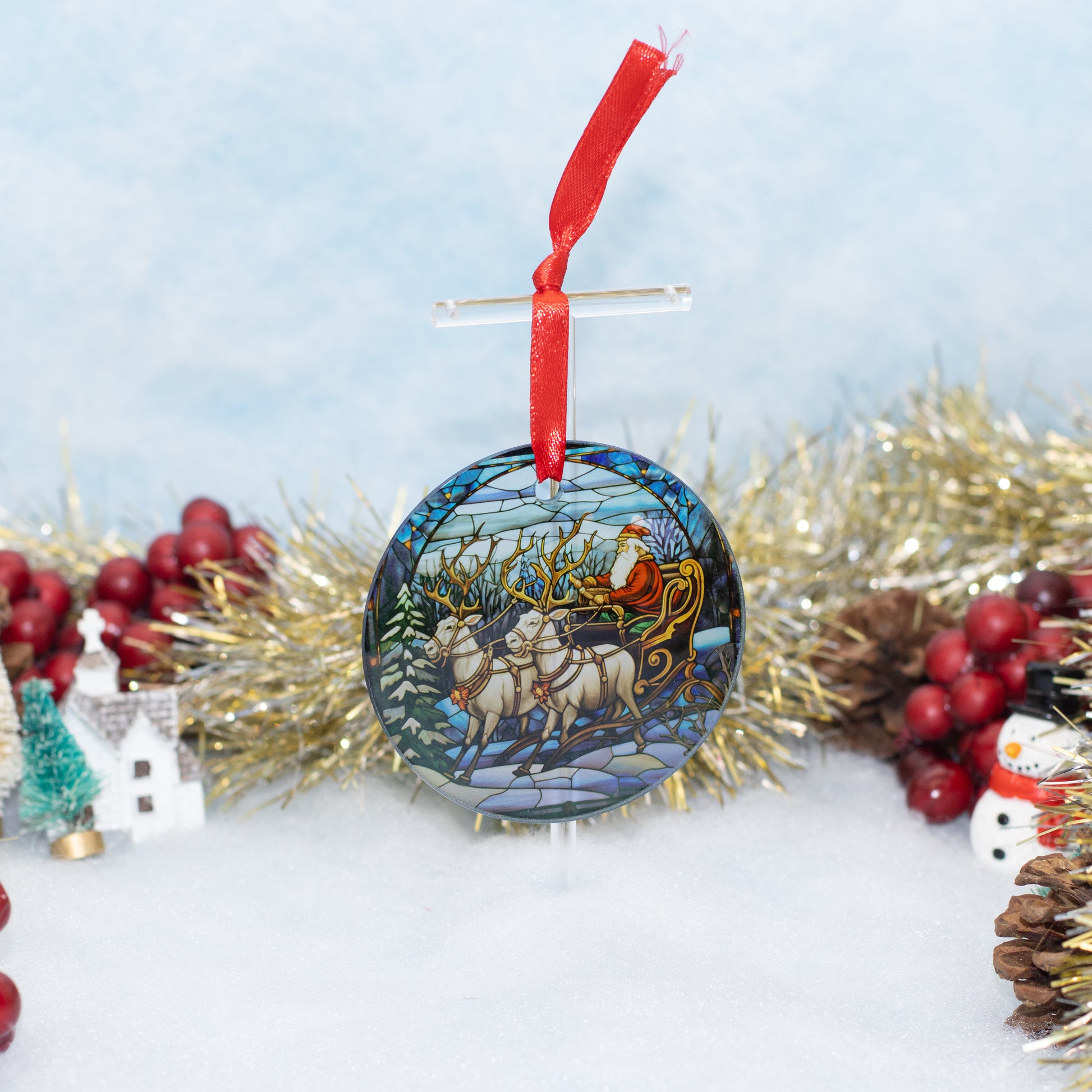A round glass Christmas ornament featuring Santa Claus in his sleigh pulled by reindeer, hanging from a red ribbon against a snowy winter scene with tinsel and berries.
