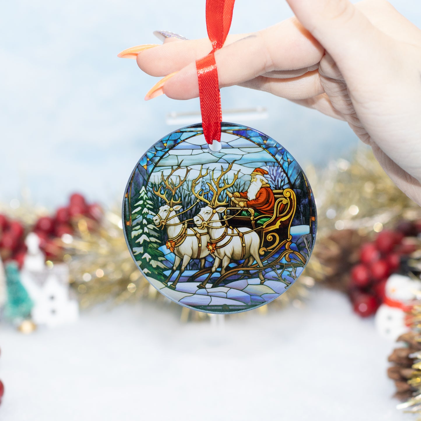A hand holds a round glass Christmas ornament featuring a stained-glass-style winter scene with Santa in a sleigh pulled by reindeer. The ornament has a red hanging ribbon, and blurred holiday decorations appear in the background.