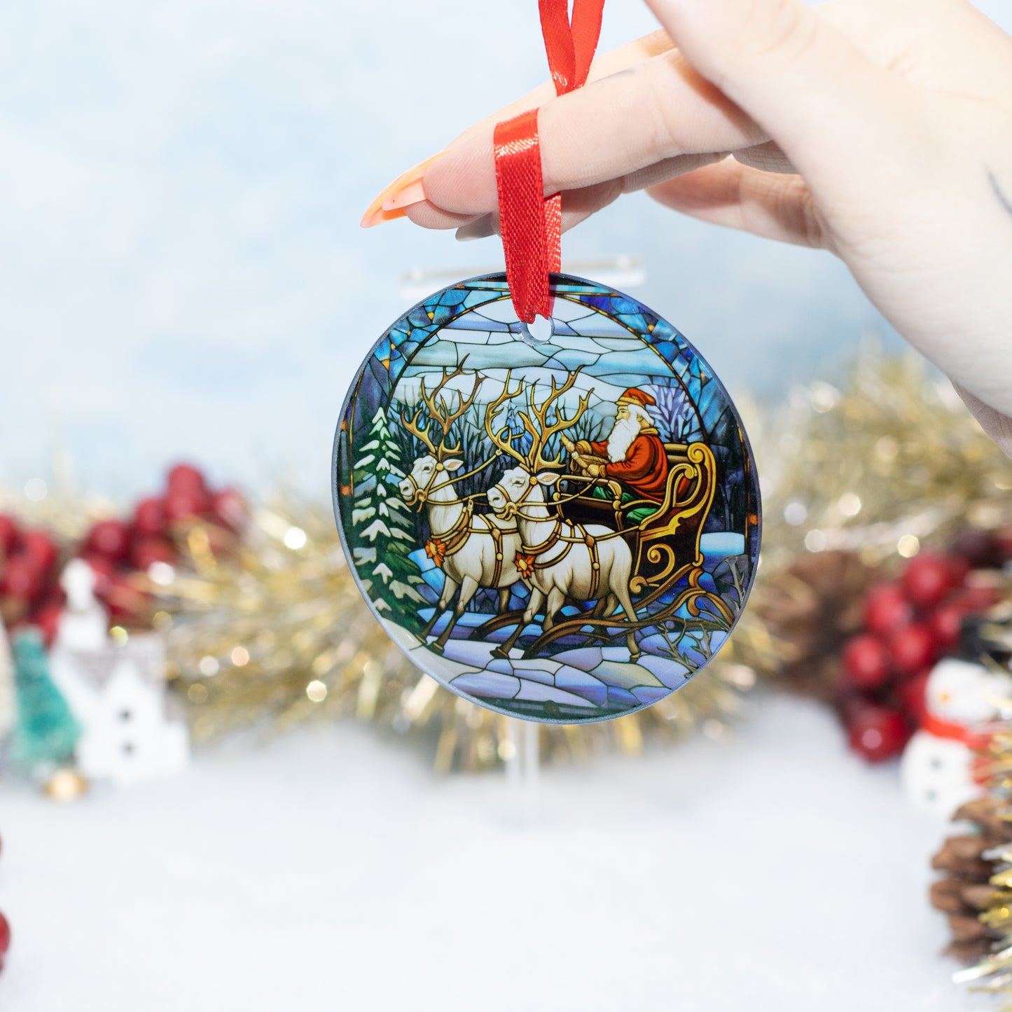 A hand holds a round glass Christmas ornament featuring a stained-glass style illustration of Santa Claus in a sleigh pulled by white deer, with a red ribbon hanging from the top. The background is softly blurred with festive decorations.
