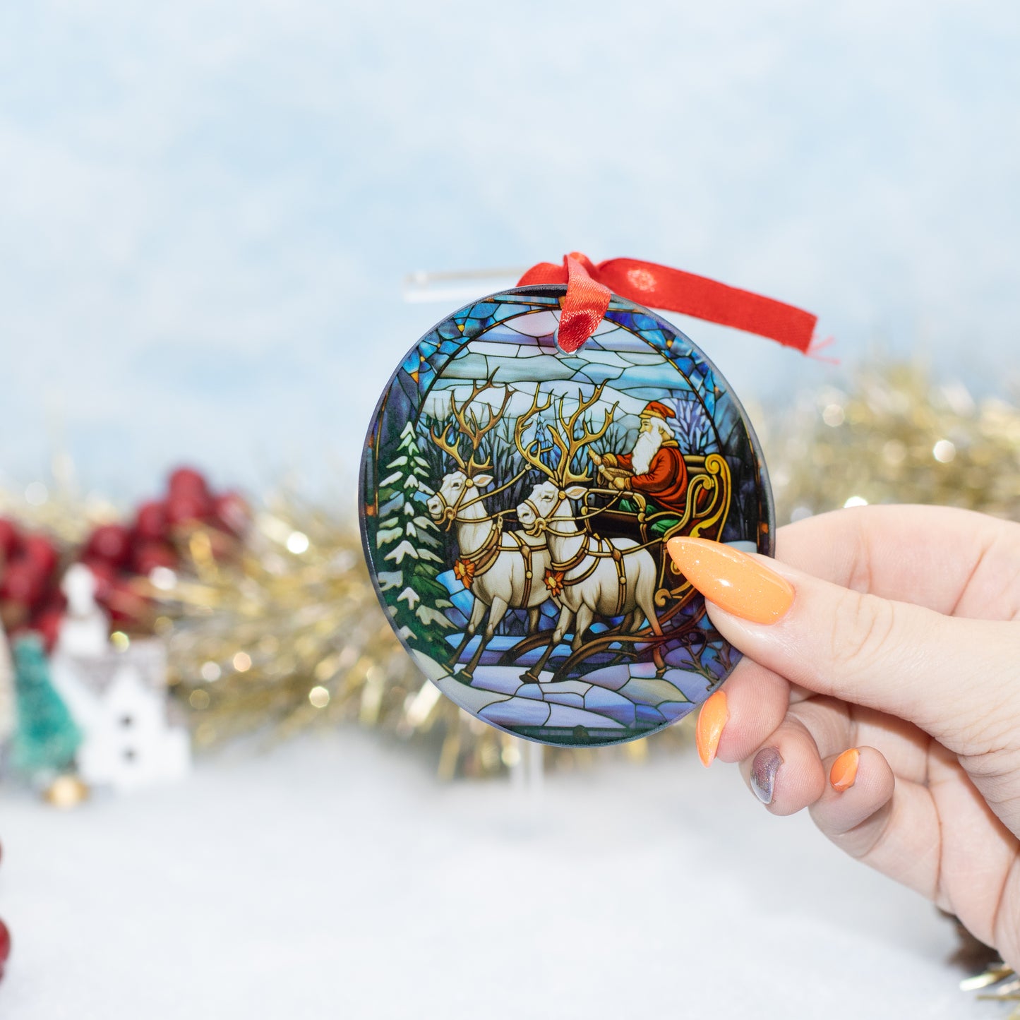 A hand holds a round stained-glass style Christmas ornament featuring Santa Claus in a sleigh with reindeer. The ornament has a red ribbon hanging from the top and is photographed against a softly blurred festive background with gold tinsel and red decorations.