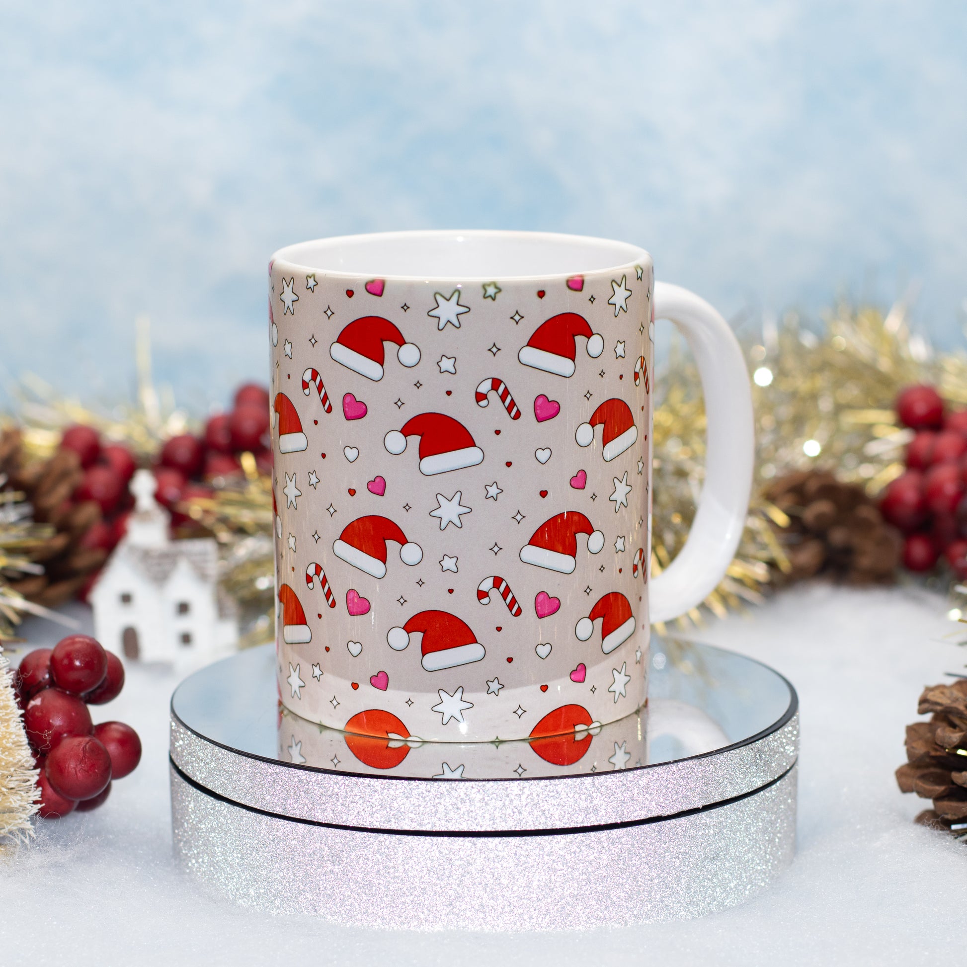 A white ceramic mug with a festive Christmas print featuring Santa hats, candy canes, hearts, and stars on a beige background, displayed on a round silver glitter pedestal with holiday decorations in the background.