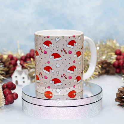A white ceramic mug with a festive Christmas print featuring Santa hats, candy canes, hearts, and stars on a beige background, displayed on a round silver glitter pedestal with holiday decorations in the background.