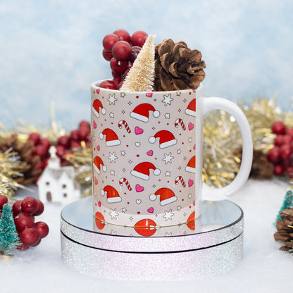 A white ceramic mug with a Christmas pattern of Santa hats, candy canes, and hearts, filled with holiday decorations like a pinecone and miniature tree, displayed on a glittery silver pedestal.