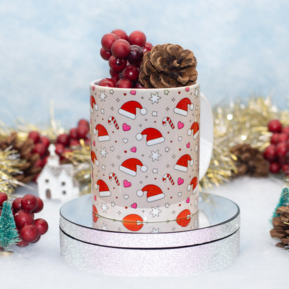 A festive ceramic mug with a Christmas print of Santa hats, candy canes, and hearts, styled with red berries and a pinecone on a mirrored silver stand against a soft blue holiday background.