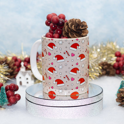 A white ceramic mug with a Christmas Santa hat pattern, filled with red berries and a pinecone, displayed on a round silver glitter pedestal against a festive holiday background.