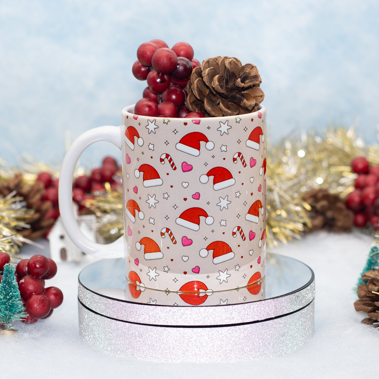 A white ceramic mug decorated with a festive Christmas pattern of Santa hats, candy canes, hearts, and stars, filled with red berries and a pinecone, displayed on a round silver glitter pedestal.
