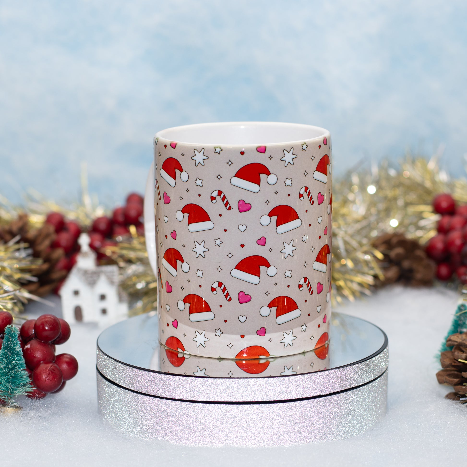 A ceramic mug with a Christmas pattern of Santa hats, candy canes, hearts, and stars on a beige background, displayed on a round silver glitter stand with festive holiday decorations in the background.