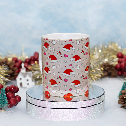 A ceramic mug with a Christmas pattern of Santa hats, candy canes, hearts, and stars on a beige background, displayed on a round silver glitter stand with festive holiday decorations in the background.