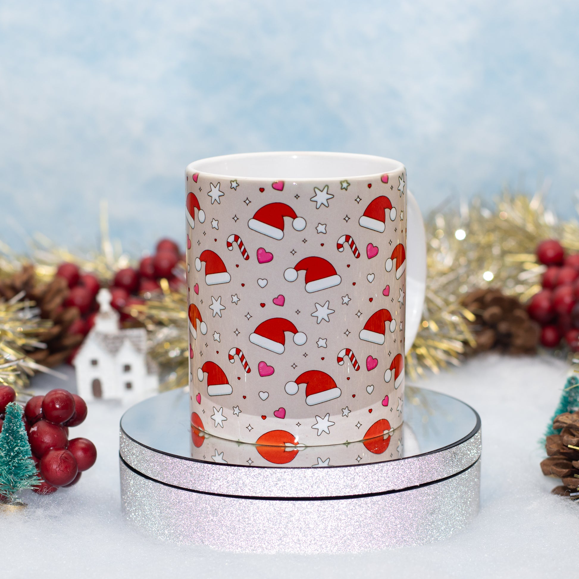 A cylindrical ceramic mug with a festive Christmas print featuring Santa hats, candy canes, hearts, and stars on a light beige background, displayed on a round silver glitter pedestal with holiday decorations in the background.