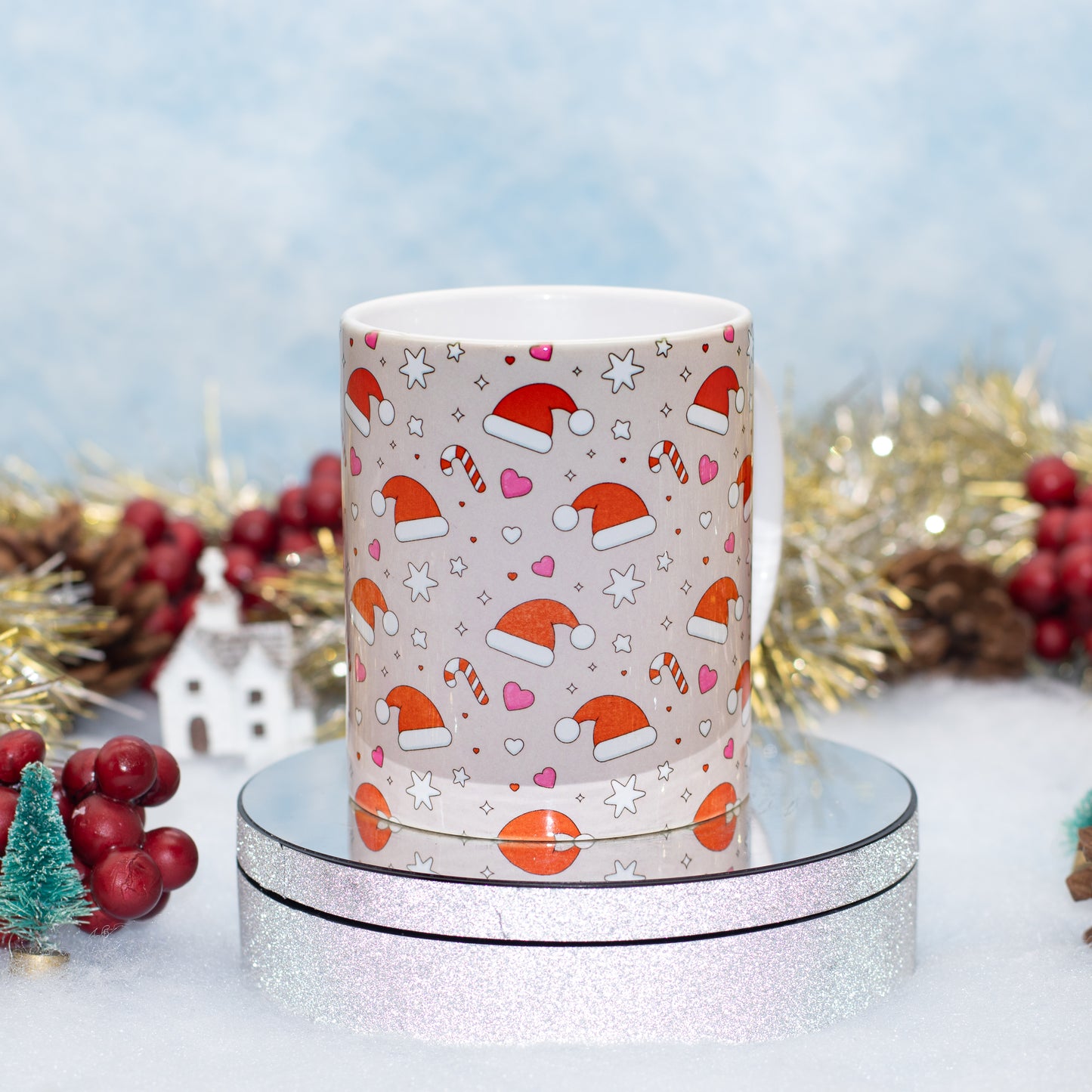 A white ceramic mug with a festive Christmas pattern of Santa hats, candy canes, hearts, and stars, displayed on a round silver glitter stand against a decorated holiday background.