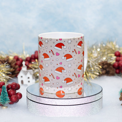 A white ceramic mug with a festive Christmas pattern of Santa hats, candy canes, hearts, and stars, displayed on a round silver glitter stand against a decorated holiday background.