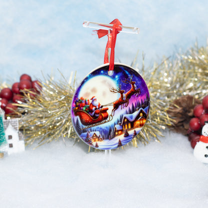 A round Christmas ornament featuring a colorful illustration of Santa Claus in his sleigh under a full moon, hanging from a red ribbon on a clear stand against a snowy, decorated holiday background.