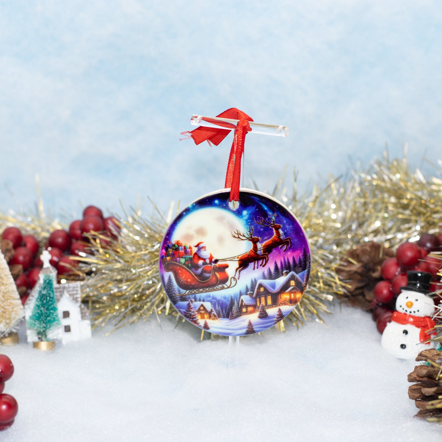 A round Christmas ornament featuring a colorful illustration of Santa Claus in his sleigh under a full moon, surrounded by gold tinsel, red berries, pinecones, and small holiday decorations on a snowy white surface.