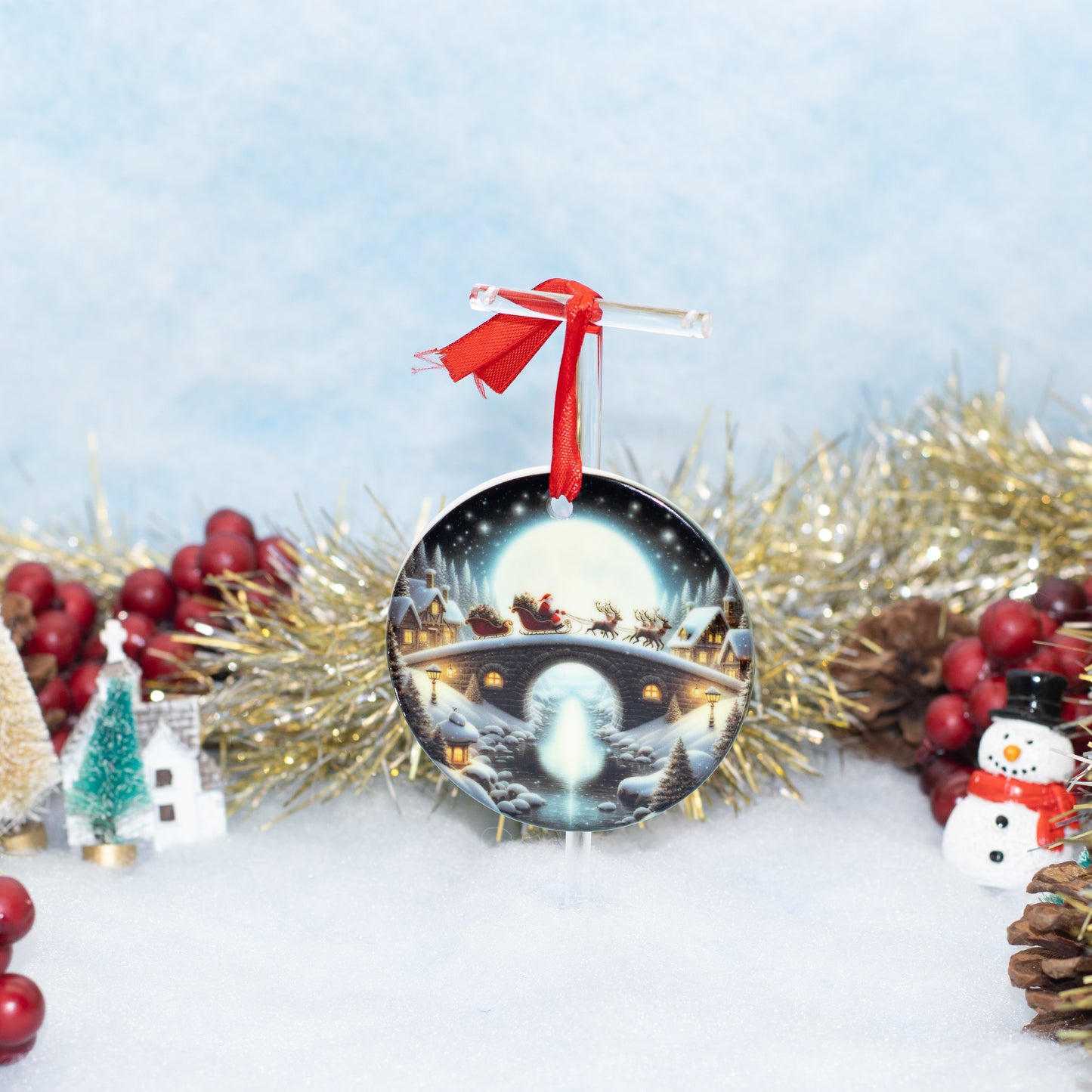 A round Christmas ornament featuring a snowy night scene with Santa's sleigh under a full moon, hanging from a red ribbon, styled with gold tinsel, pinecones, red berries, and small holiday decorations on a snowy white surface.