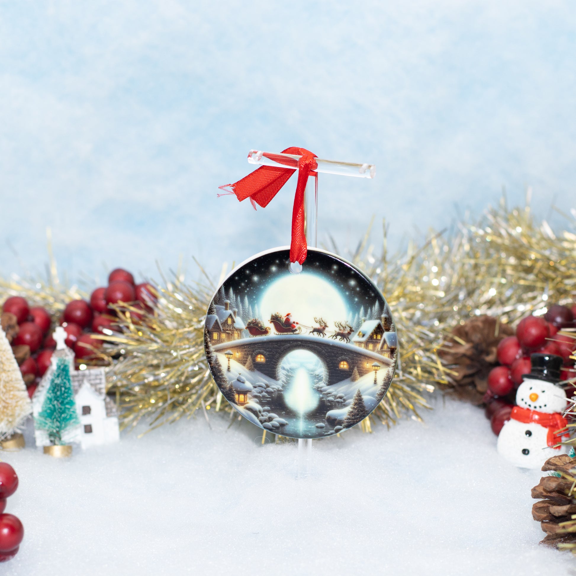 A round Christmas ornament featuring a snowy night scene with Santa's sleigh under a full moon, hanging from a red ribbon, styled with gold tinsel, pinecones, red berries, and small holiday decorations on a snowy white surface.
