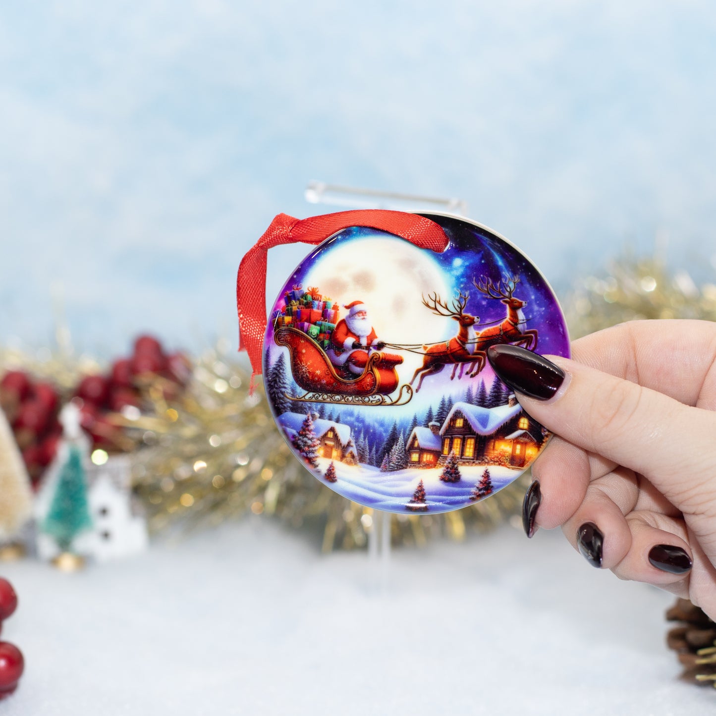 A hand holds a round Christmas ornament featuring Santa Claus in a red sleigh under a full moon, with snowy houses and reindeer, against a softly blurred festive background.