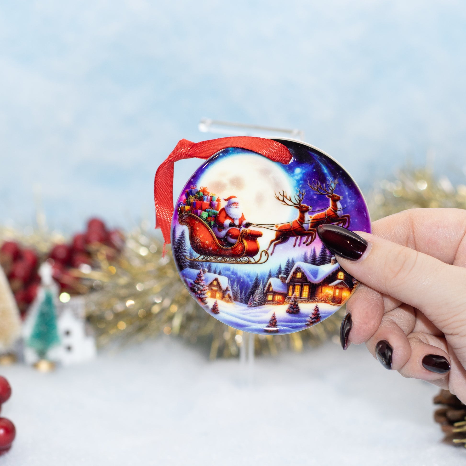 A hand holds a round Christmas ornament featuring Santa Claus in a red sleigh under a full moon, with snowy houses and reindeer, against a softly blurred festive background.