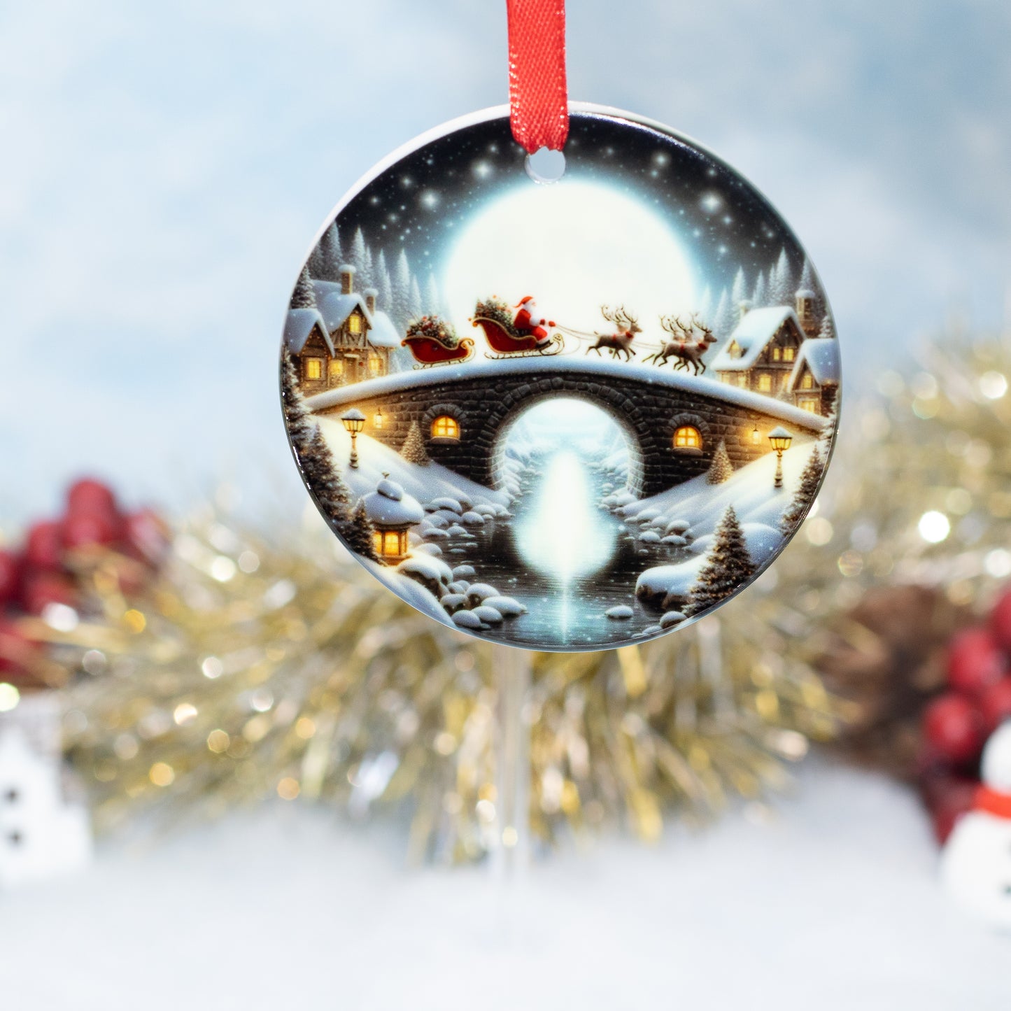 A round Christmas ornament featuring a festive winter village scene with Santa Claus in his sleigh, snow-covered houses, evergreen trees, and a glowing bridge under a full moon. The ornament hangs from a red ribbon against a softly blurred holiday background with gold tinsel and red berries.