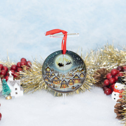 A round Christmas ornament featuring a painted winter village scene under a full moon with Santa's sleigh, hanging from a clear stand with a red ribbon. The ornament is styled on a snowy surface with gold tinsel, red berries, pinecones, a small tree, and a snowman around it.