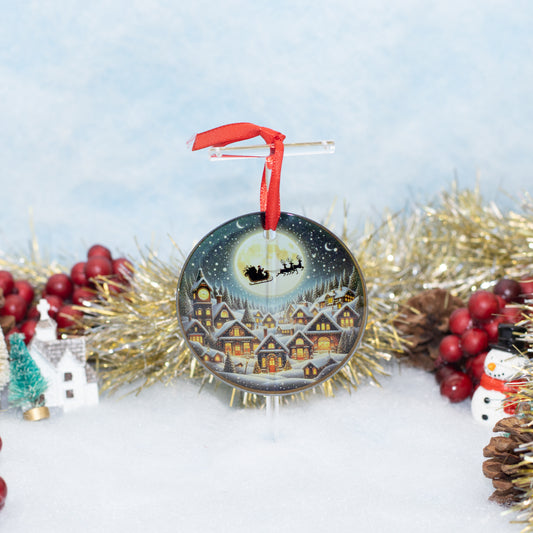 A round Christmas ornament featuring a painted winter village scene under a full moon with Santa's sleigh, hanging from a clear stand with a red ribbon. The ornament is styled on a snowy surface with gold tinsel, red berries, pinecones, a small tree, and a snowman around it.